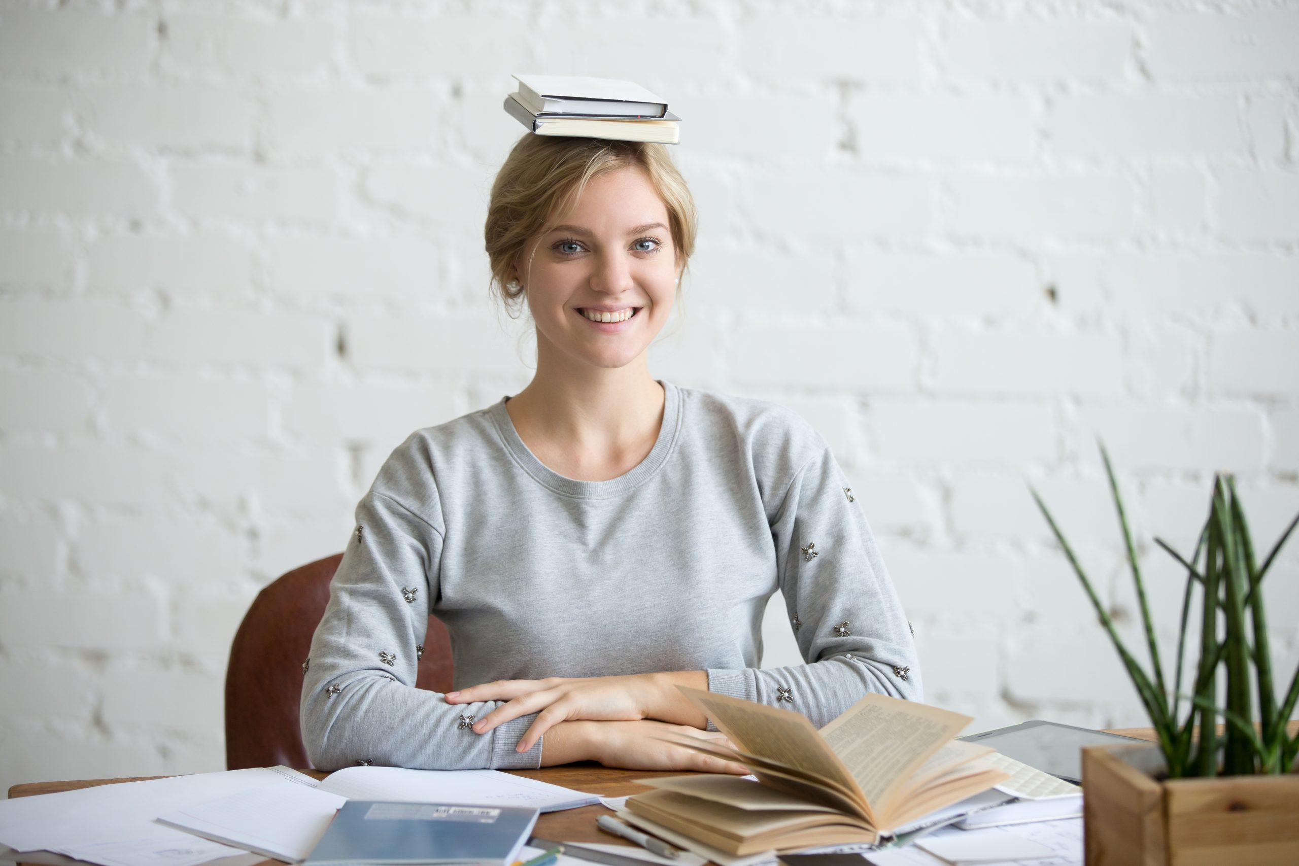 Portrait of a young smiling woman at the desk with books on her head, sitting straight, looking at the camera. Education concept photo